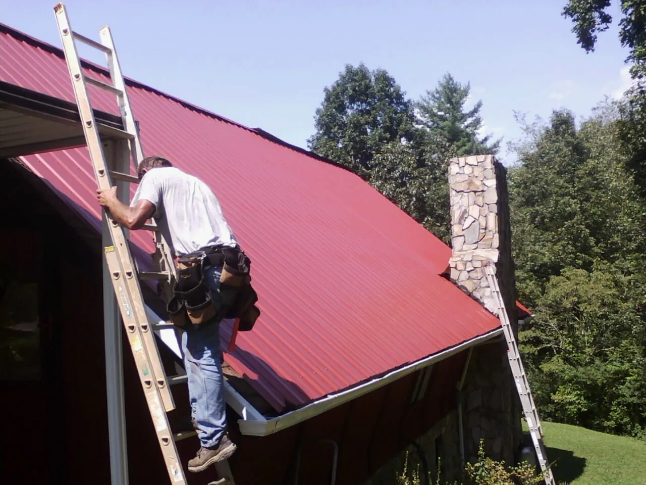 Roofer on a ladder beside a red metal roof and stone chimney — Soffit & Fascia Repair by Roofing Brothers in North Bonneville